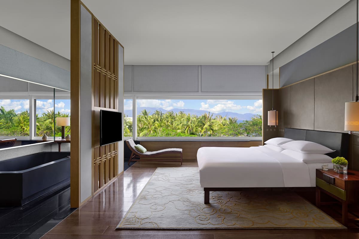 A modern hotel room with a large bed, a sitting area with a chair and a small table, a bathtub, and a large window offering a view of palm trees and mountains.