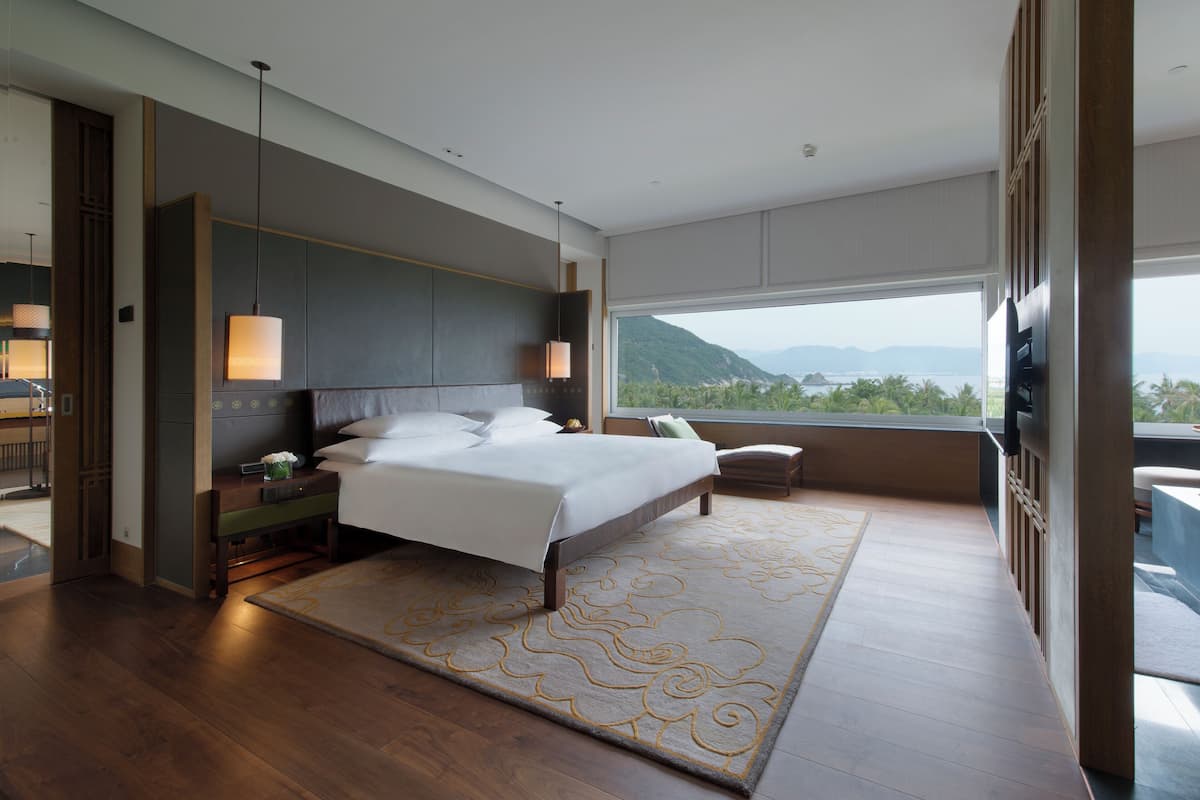 A modern bedroom with a large bed, bedside tables, a view of mountains through a large window, and a balcony.