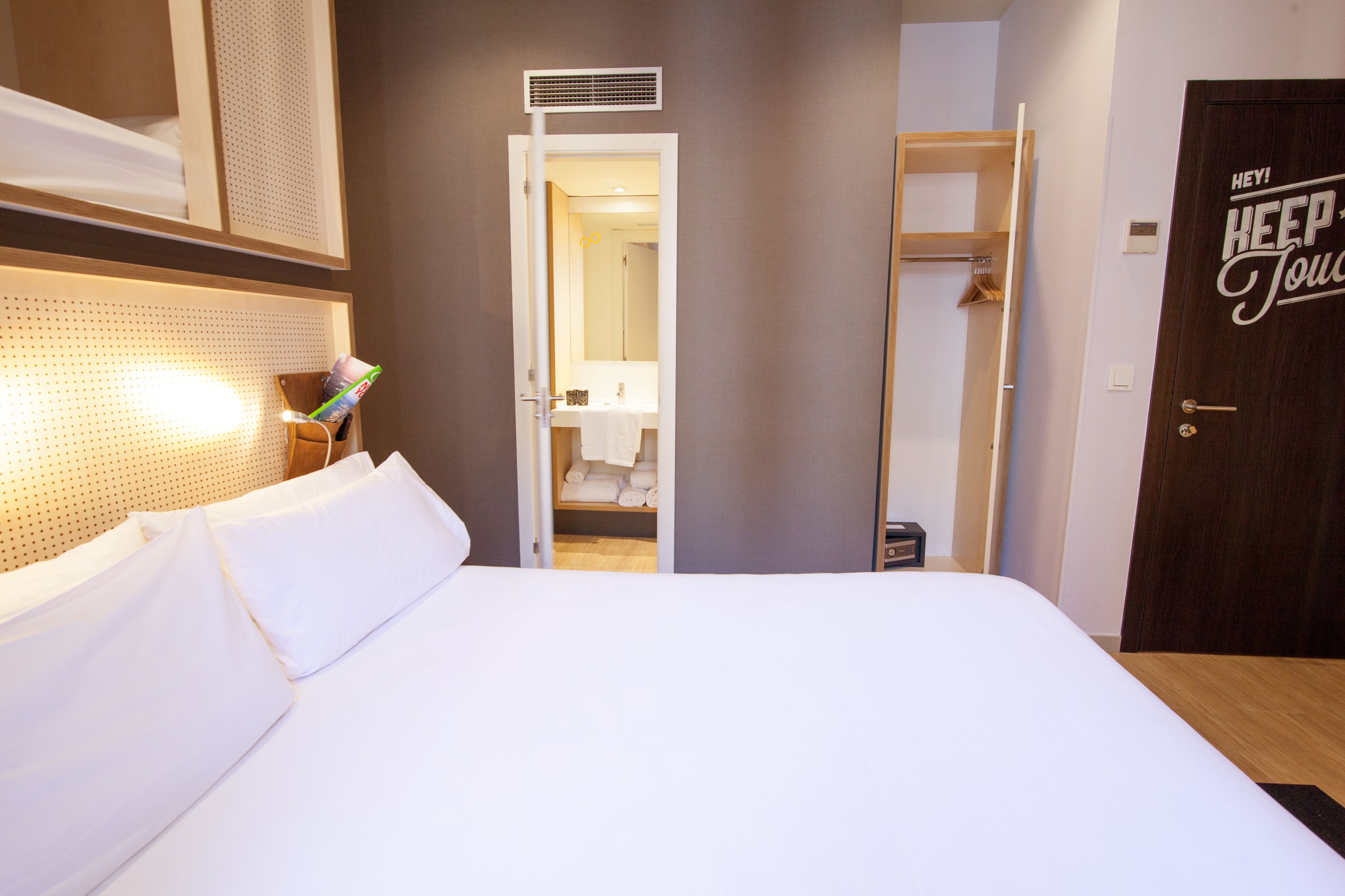 A hotel room with a neatly made bed, a bathroom visible through an open door, and a wardrobe with a 'KEEP TOUCH' sign.
