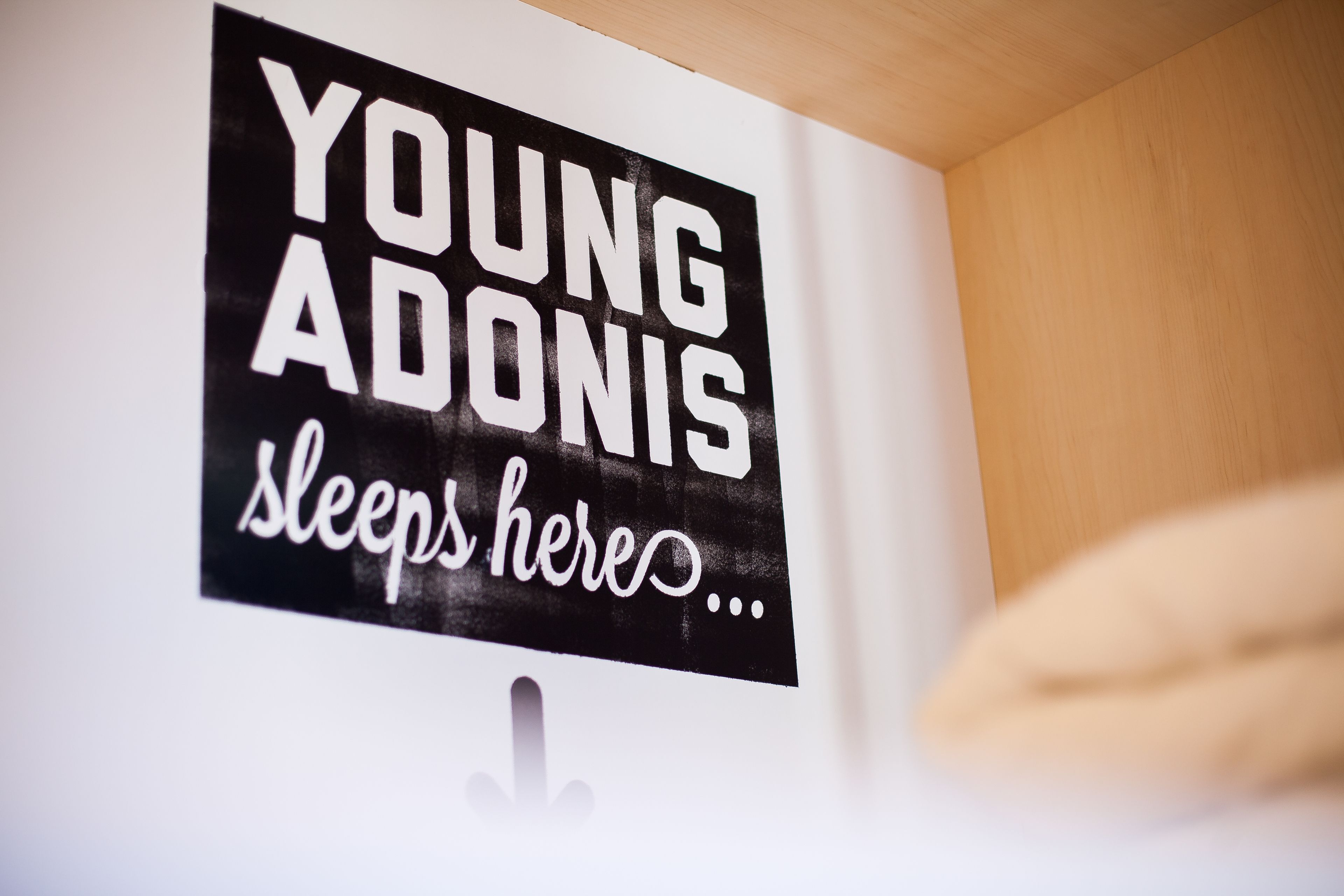 Una señal con el texto 'YOUNG ADONIS duerme aquí' en una pared.