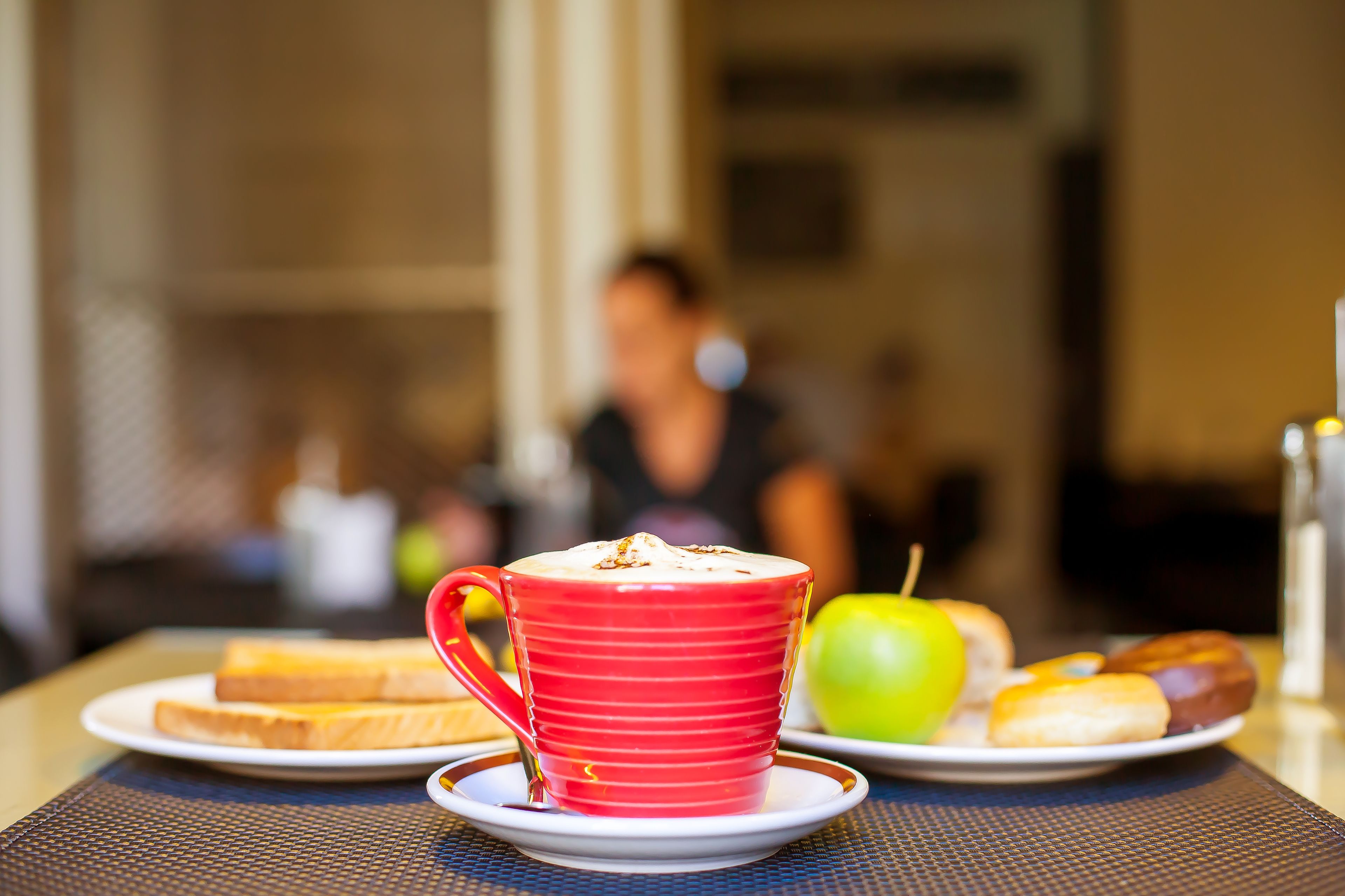 A breakfast setting with a red cup of coffee, toast, an apple, and pastries on a table.