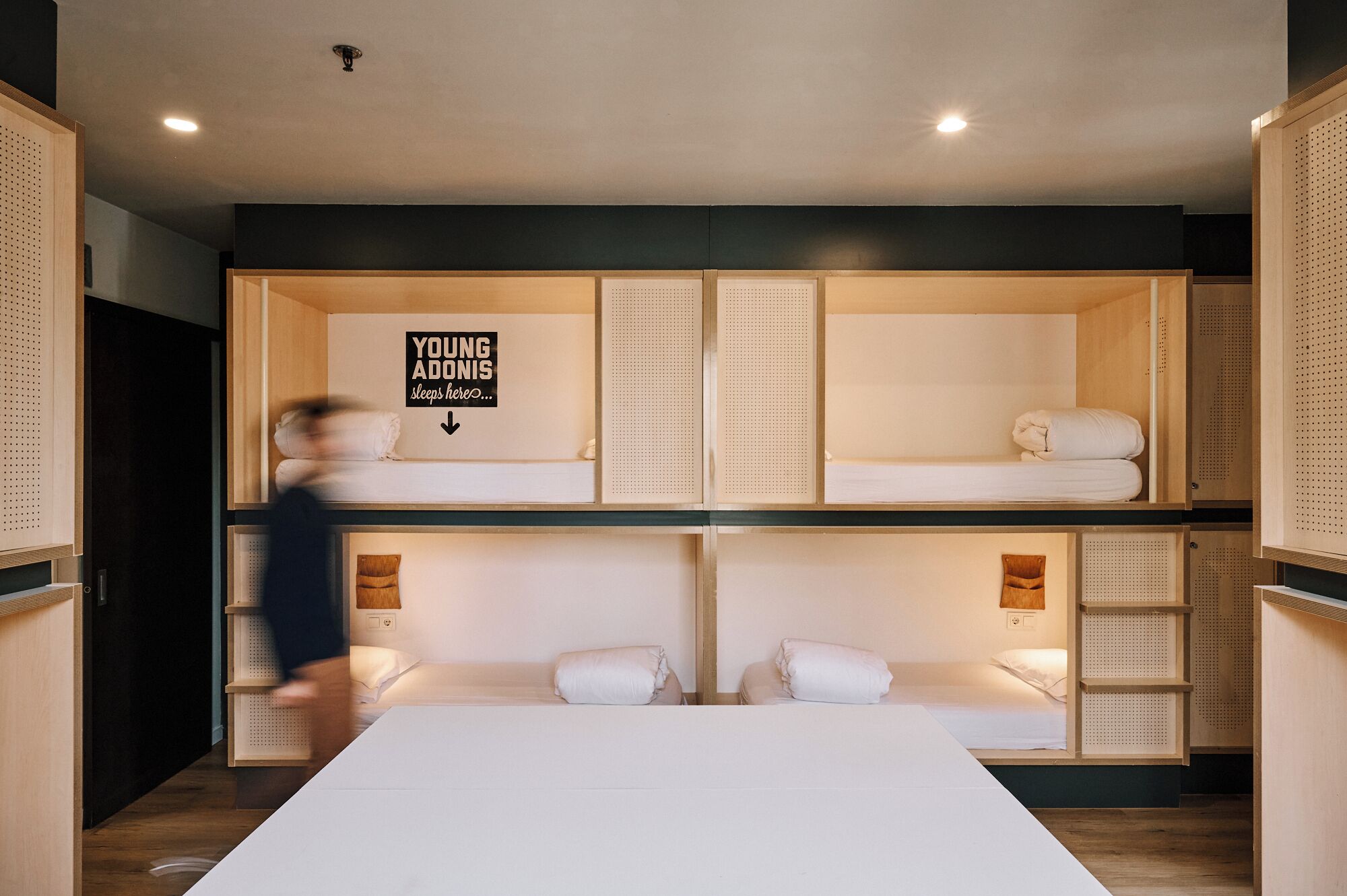 A modern, multi-level dormitory room with bunk beds, a central desk, and a sign reading "Young Adonis sleeps here."