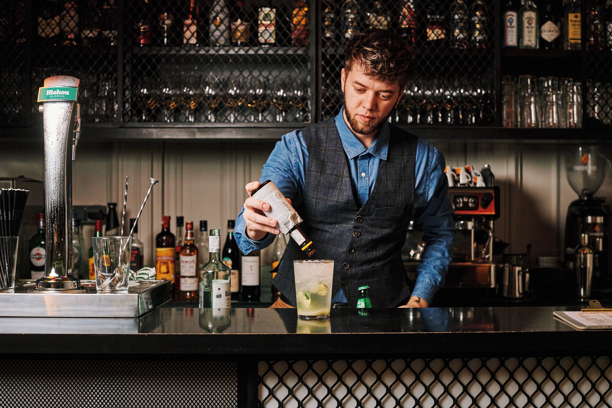 Een barman schenkt een drankje in een glas, op de achtergrond een diverse selectie flessen en glazen.