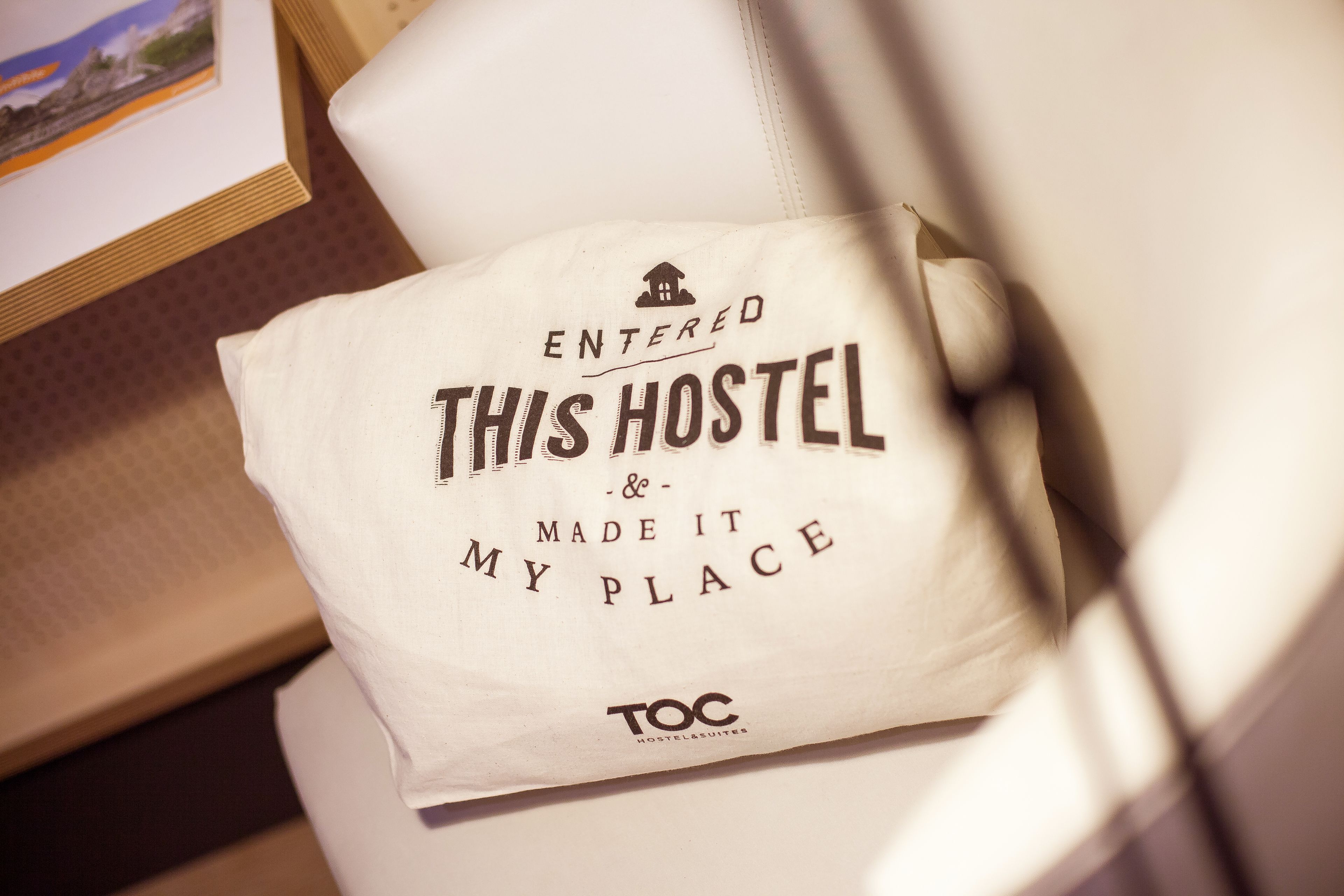 A pillow with the text "ENTERED THIS HOSTEL & MADE IT MY PLACE" and the logo "TOC HOSTEL SUITES".
