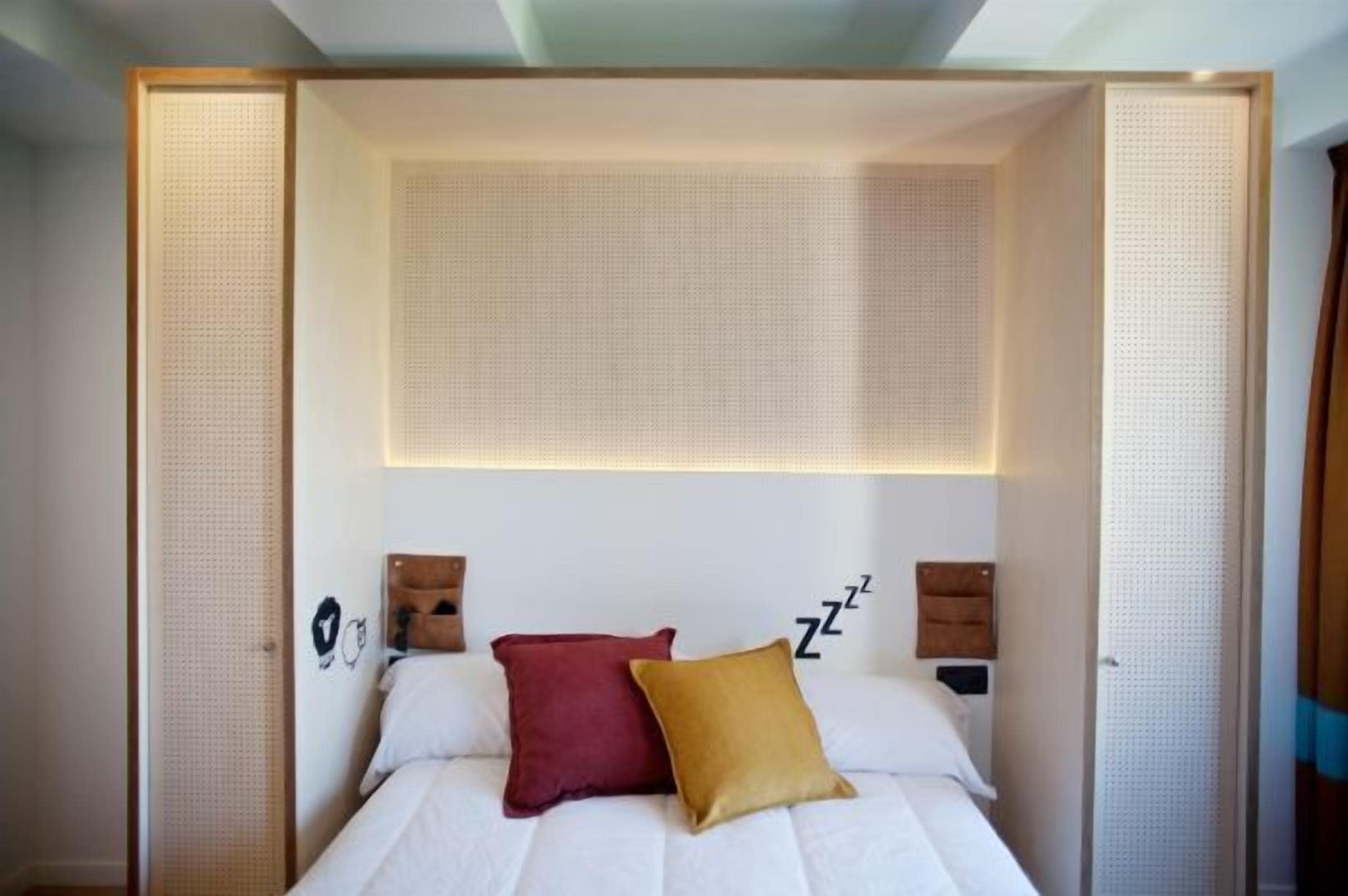A neatly made bed with two pillows, a wall-mounted clock, and a bedside table with a lamp.