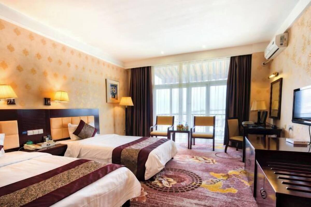 A hotel room with two beds, a desk, a TV, and a large window with curtains.