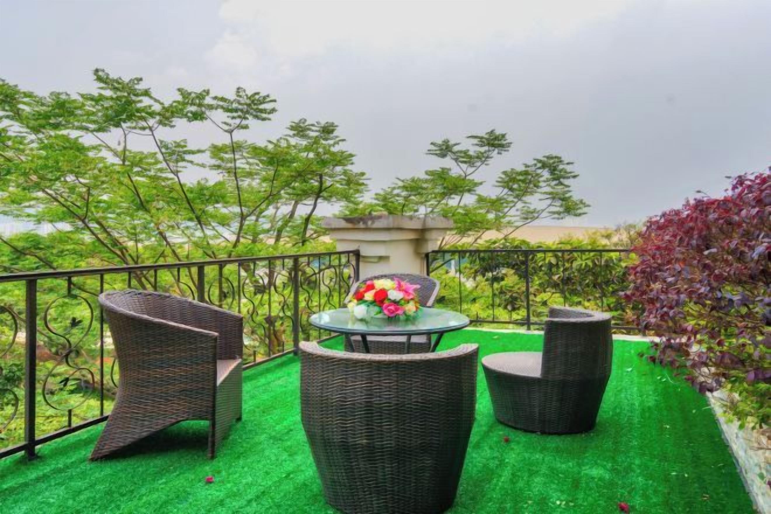 A balcony with wicker furniture and a table with flowers, surrounded by greenery and a cloudy sky.