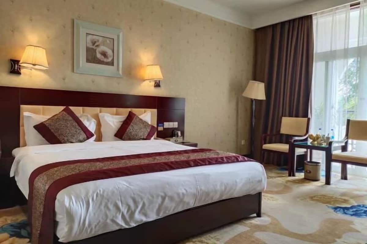 A hotel room with a large bed, two wall-mounted lamps, a framed picture, a window with curtains, and a small table with a chair.