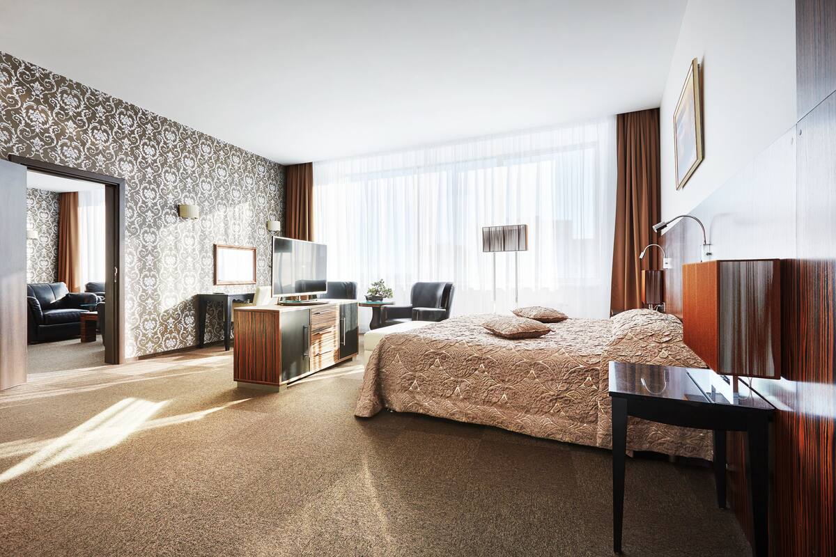 A modern hotel room with a large bed, a desk, a wardrobe, and a seating area with a sofa and chairs.