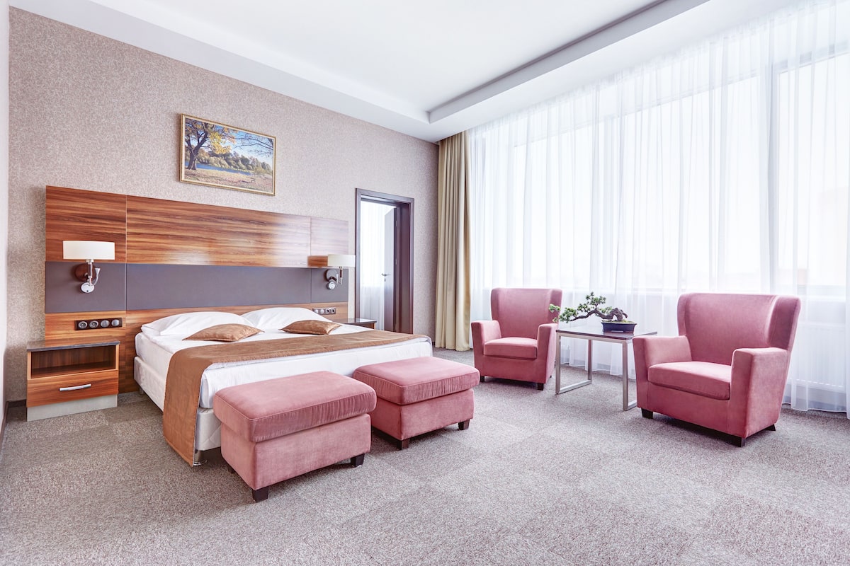 A hotel room with a large bed, two pink armchairs, a small table with a plant, and a painting on the wall.