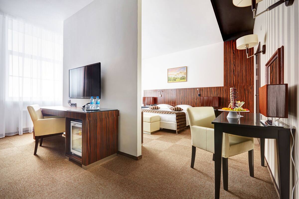 A hotel room with a TV, a desk with a computer, a dining area with a table and chairs, a bed, and a painting on the wall.
