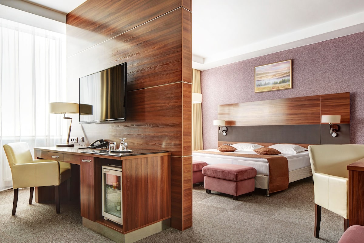 A hotel room with a wooden desk, a bed with a headboard, a television, and a painting on the wall.