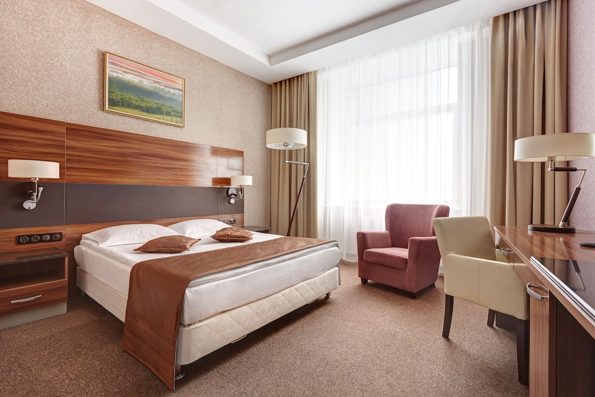A hotel room with a bed, a desk, a chair, a lamp, and a painting on the wall.