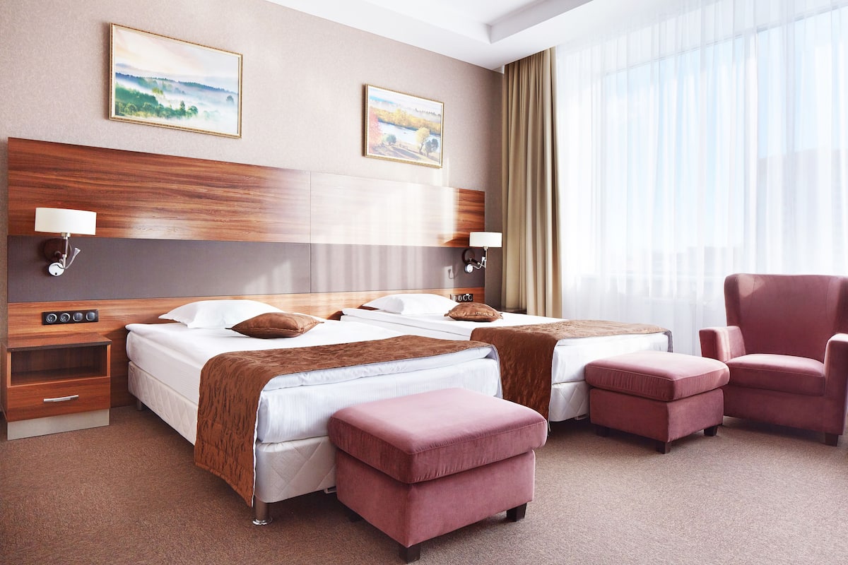 A hotel room with two beds, a wooden headboard, a red armchair, a brown ottoman, and two wall-mounted lamps.