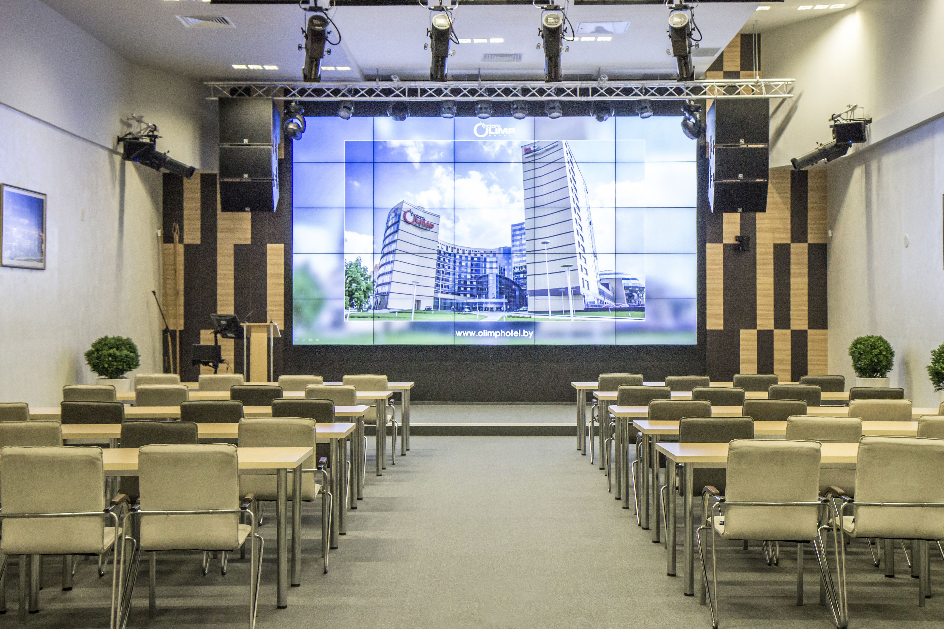 A conference room with a large screen displaying a building image, arranged seating, and a stage with lighting equipment.
