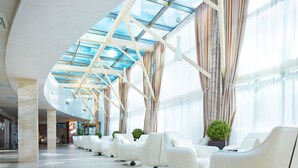 A spacious lobby with a modern design, featuring a row of white chairs, a glass ceiling, and large windows with curtains.