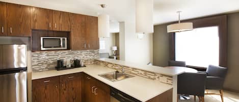 Una cocina moderna con armarios de madera, un refrigerador de acero inoxidable, un microondas y un lavavajillas. Hay un lavamanos con un amplio ventanal encima, y un comedor con sillas y una mesa.