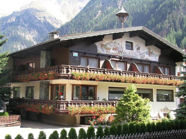 A traditional chalet with a balcony adorned with flowers, a mountain backdrop, and a sign reading "Carinthia".