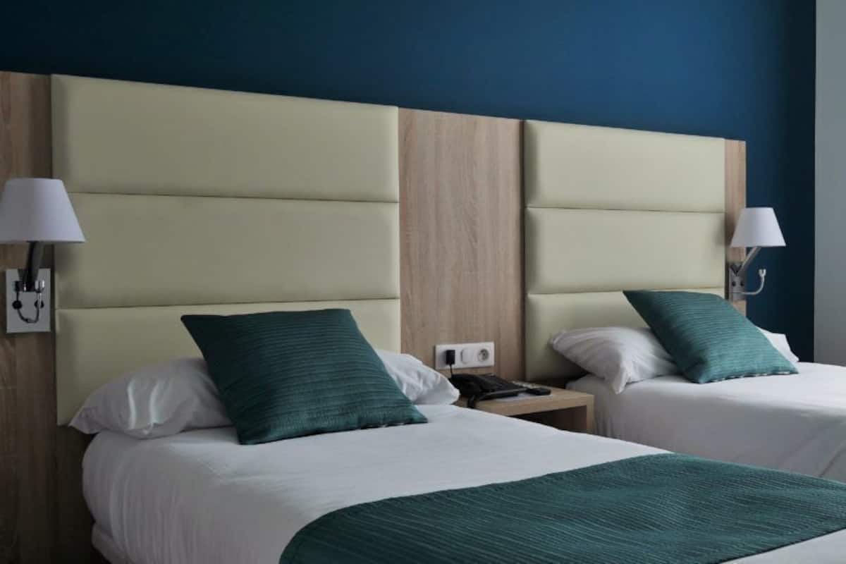 A hotel room with two beds, each with a green bedspread and white pillows. There are bedside lamps on either side of the beds, and a nightstand with a phone between them.