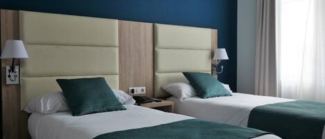 A hotel room with two beds, each with a green bedspread and white pillows. There are bedside lamps on either side of the beds, and a nightstand with a phone between them.