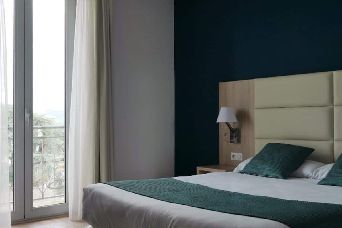 A hotel room with a large bed, a nightstand, a lamp, and a view of the outdoors through a window.