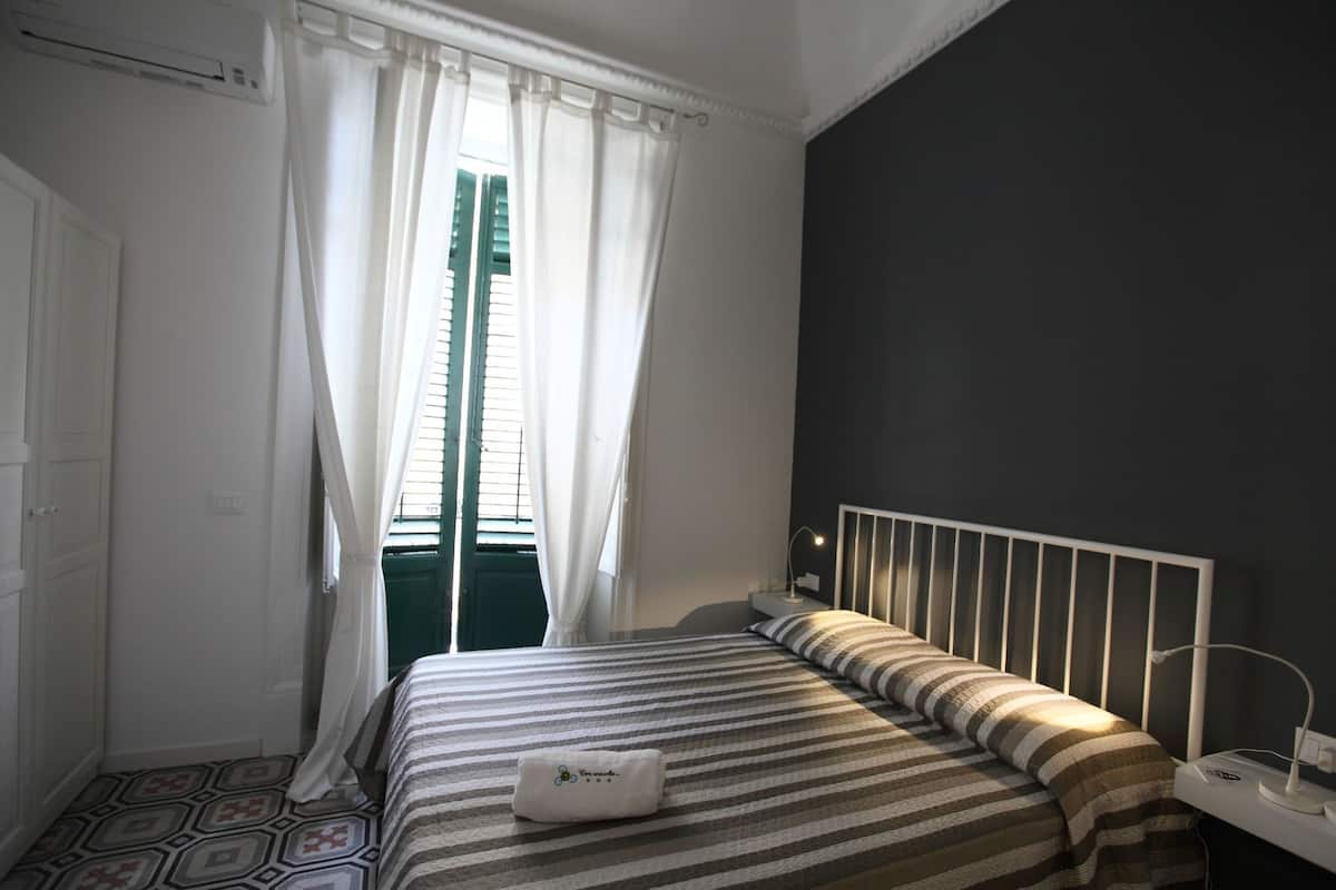 Un dormitorio con una cama, una mesita de noche con lámpara, una ventana con cortinas y un radiador.