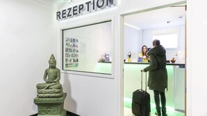 A reception area with a Buddha statue, a framed informational display, and a counter where a person is interacting with a staff member. - NXT PARK Hamburg by ANS (Hamburg)