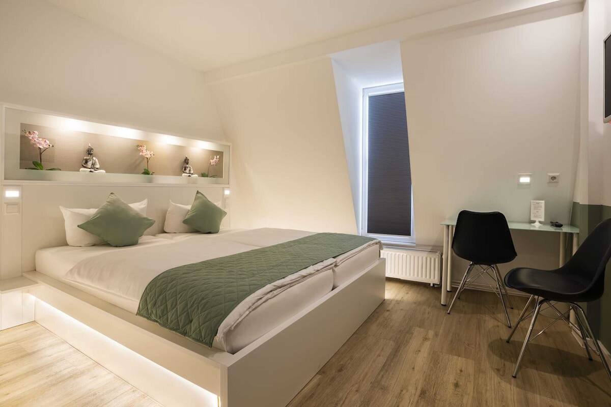 Quarto moderno com uma cama grande, uma escrivaninha e uma cadeira. Há uma janela com persianas, um radiador e uma prateleira com objetos decorativos.