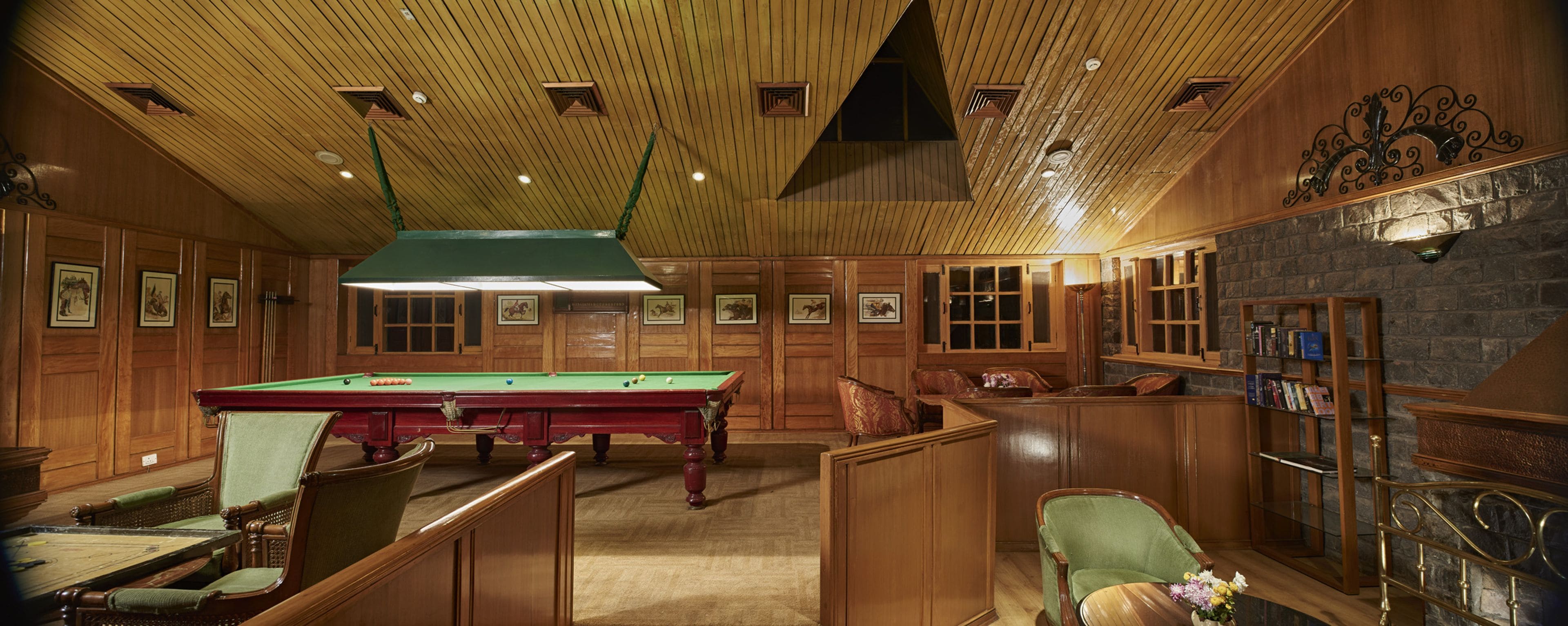 A billiards room with a pool table, green cushioned chairs, wooden paneling, and a stone fireplace.