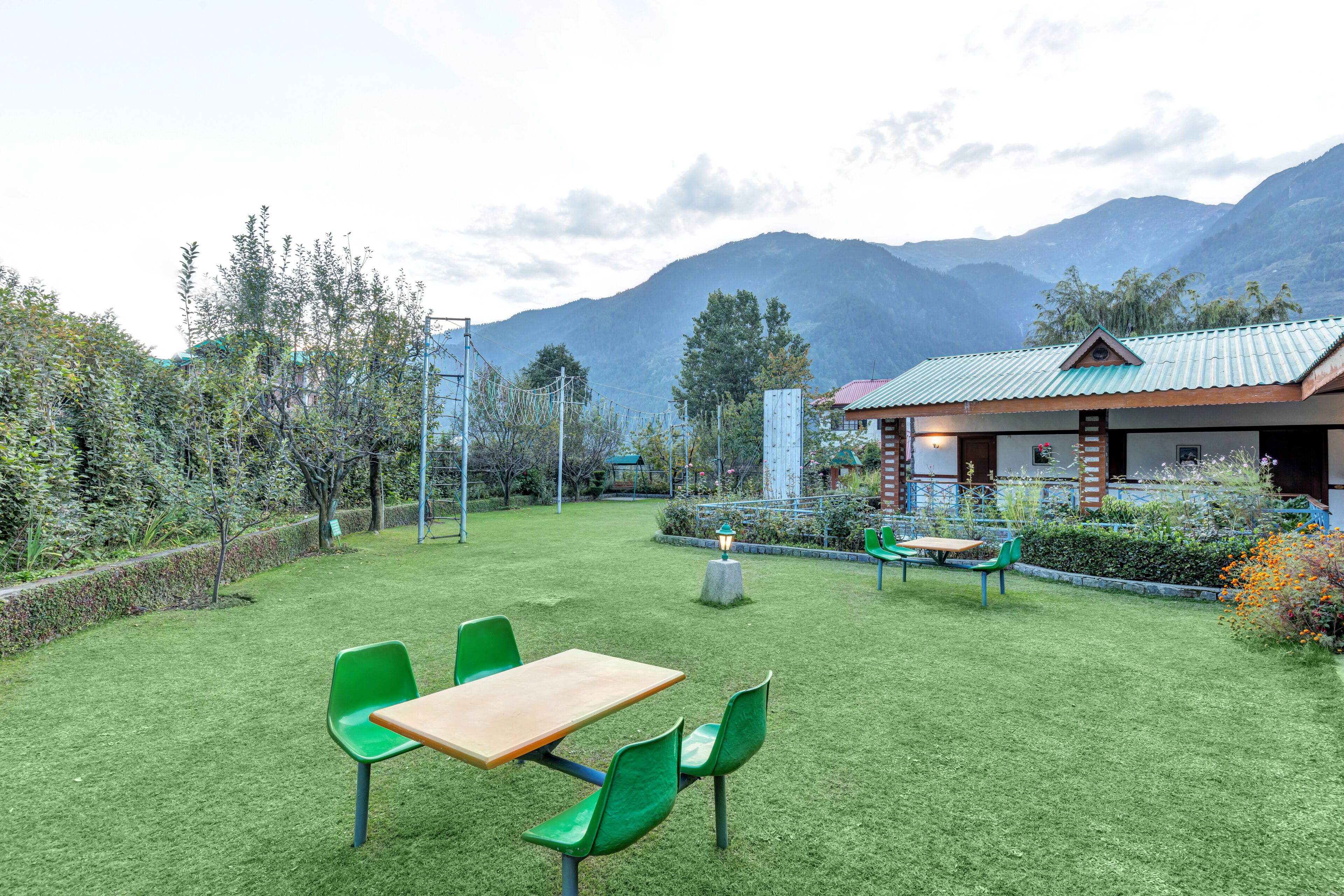 A grassy outdoor area with a wooden table and green chairs, a small building with a red roof, and mountains in the background.