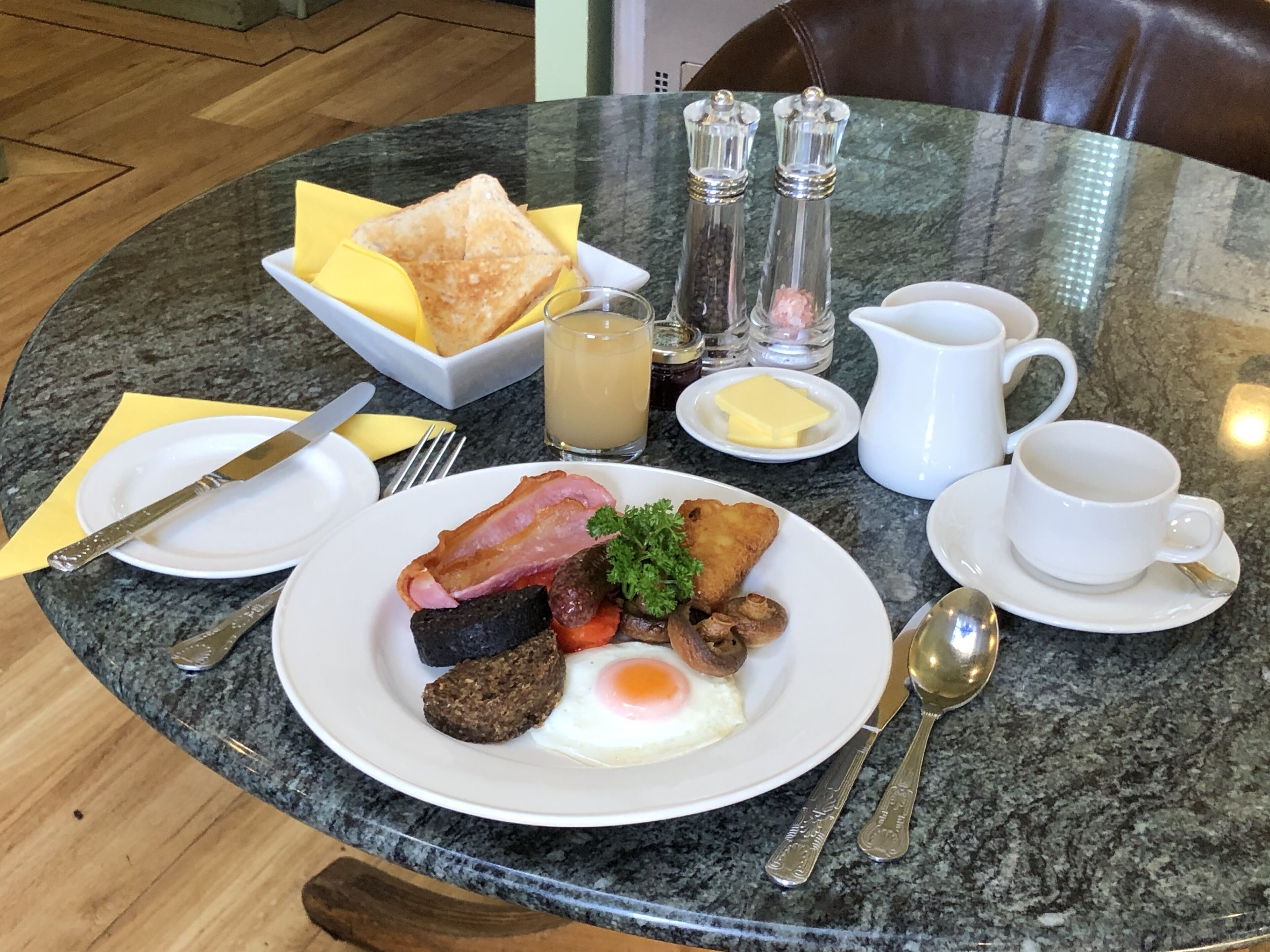 Une table de petit-déjeuner avec une assiette d’œufs, de bacon, de champignons et de pain grillé, accompagnée de beurre, de confiture et d’un verre de jus de fruits.
