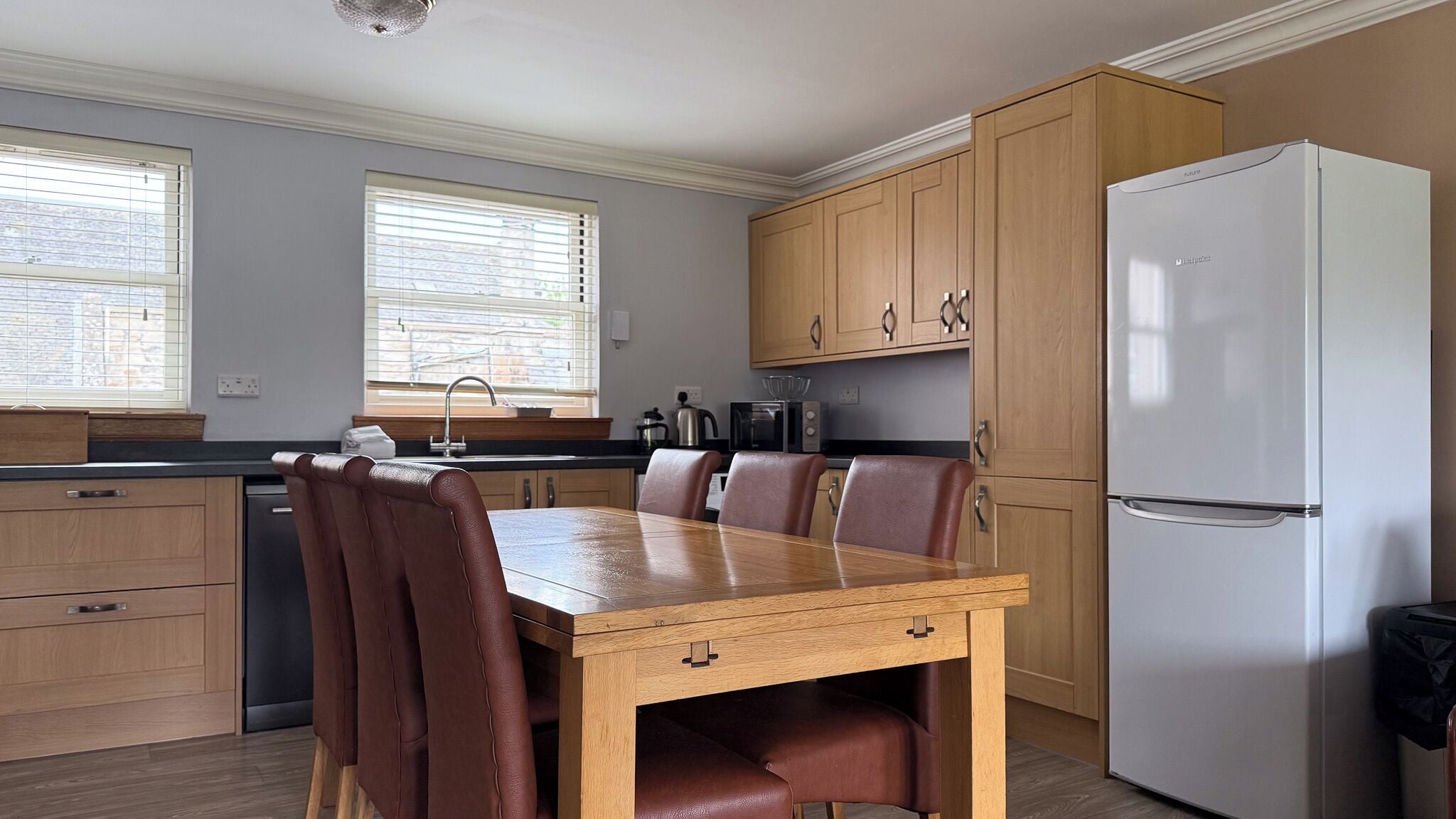 A modern kitchen with wooden cabinets, a white refrigerator, a dining table with chairs, and a window with blinds.