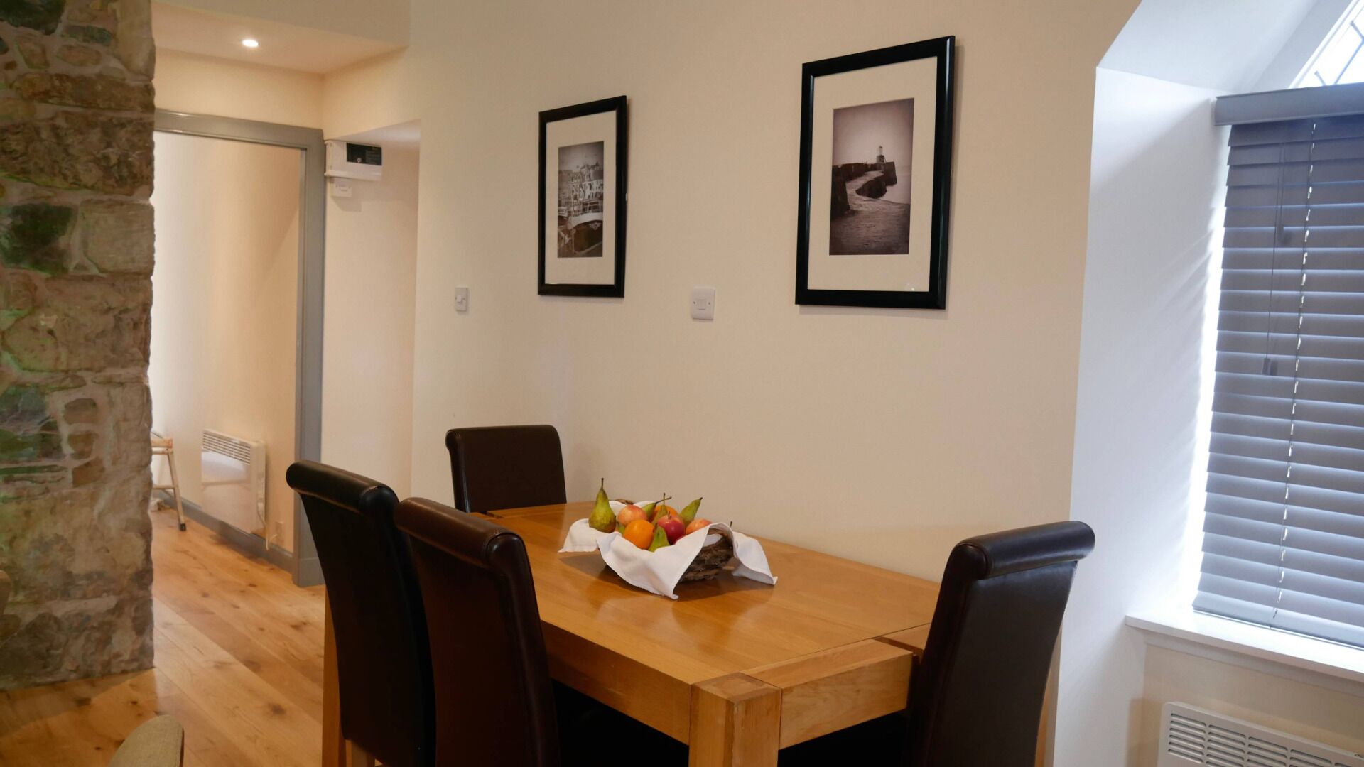 A dining area with a wooden table, four chairs, and a fruit basket. There are two framed pictures on the wall.