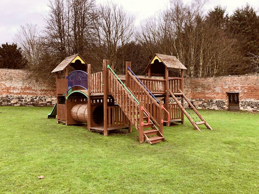 A wooden play structure with multiple levels and slides, set against a brick wall and surrounded by trees.
