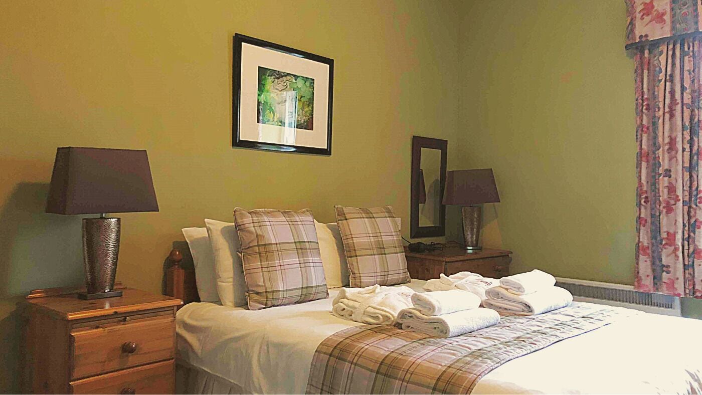 A hotel room with a bed, two bedside tables, a nightstand, a framed picture on the wall, and a window with curtains.