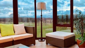 A room with a wicker sofa, a small ottoman, a floor lamp, and a large window offering a view of a grassy field and trees. - Kilconquhar Castle Estate (Leven)