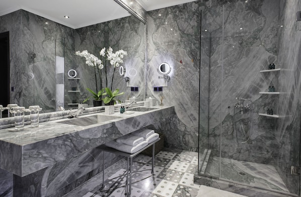 Un baño moderno con paredes de mármol, un espejo grande, dos lavamanos, una ducha y un banco.