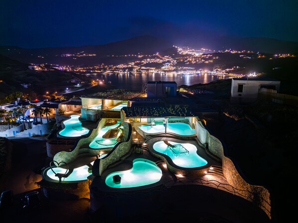A resort with multiple hot tubs illuminated at night, overlooking a coastal town and a body of water. - Agalia Luxury Suites (Ios)