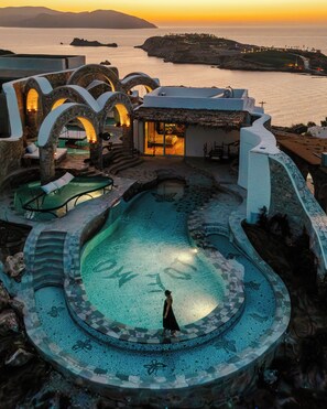 A resort with a large pool, outdoor seating, and a view of the sea at sunset. - Agalia Luxury Suites (Ios)