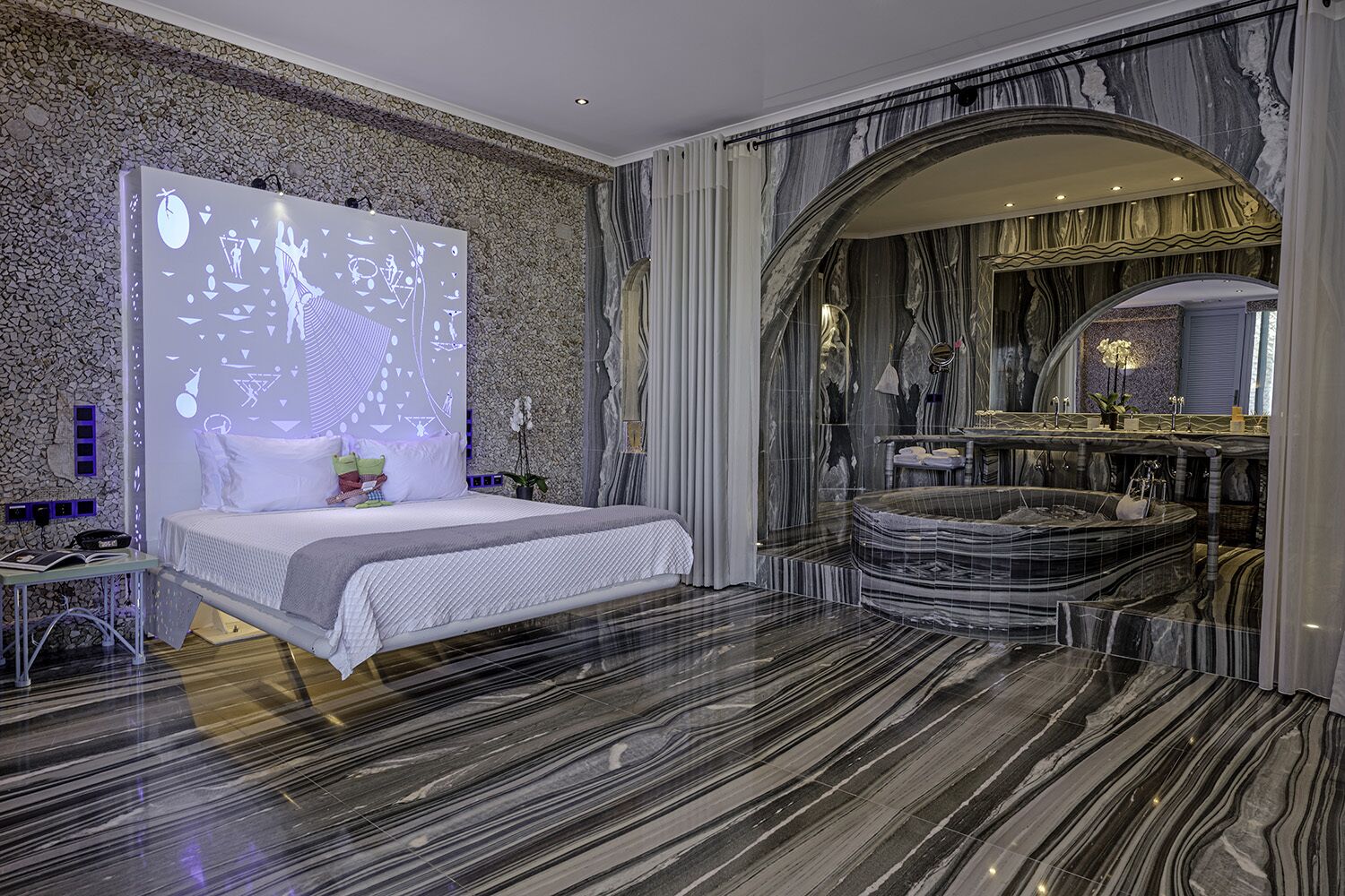 A luxurious bedroom with a large bed, a marble floor, a marble bathtub, and a stone wall.