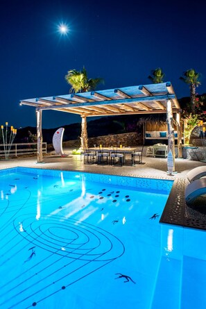 A swimming pool with underwater lights and a wooden pergola with seating. - Agalia Luxury Suites (Ios)