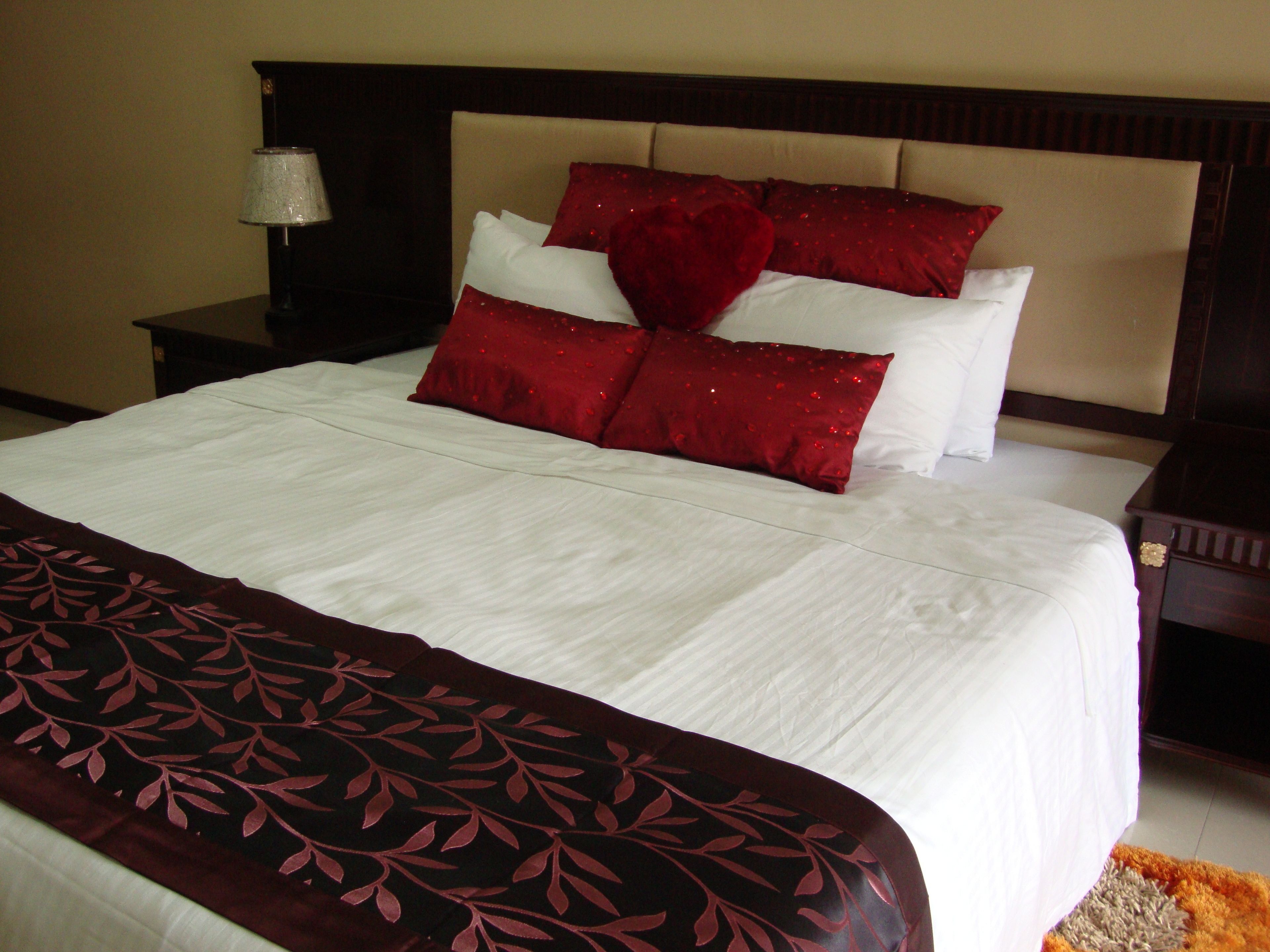 A neatly made bed with white linens and red decorative pillows, a dark wooden headboard, and a small bedside table with a lamp.