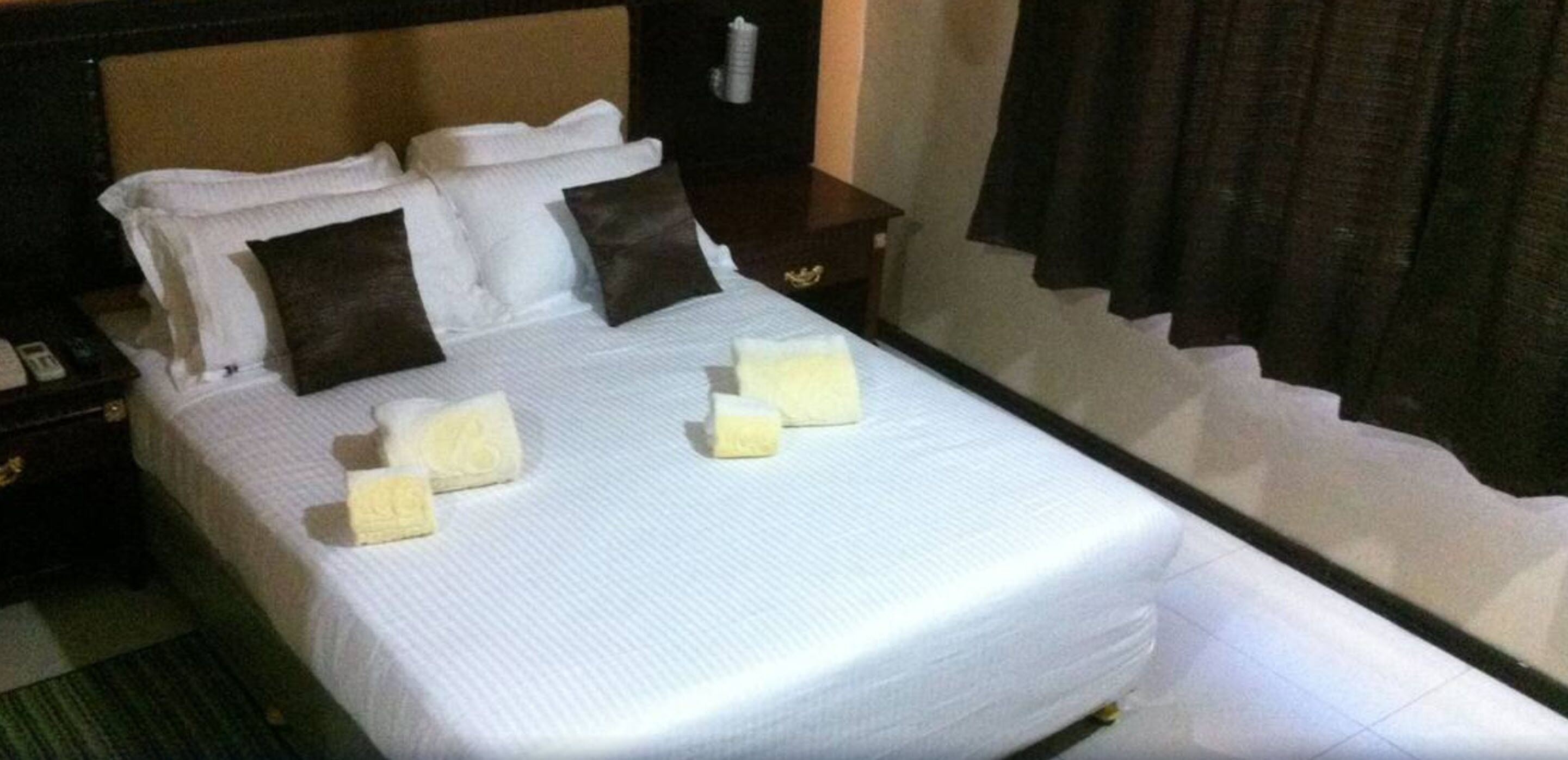 Una cama bien tendida con sábanas blancas y dos almohadas, una mesita de noche con una lámpara y una cortina oscura en la ventana.
