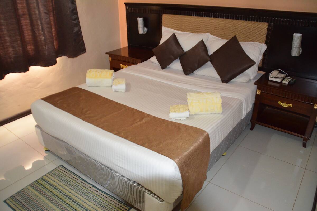 Une chambre d’hôtel avec un lit, une table de chevet, une commode, une lampe et une fenêtre avec des rideaux.