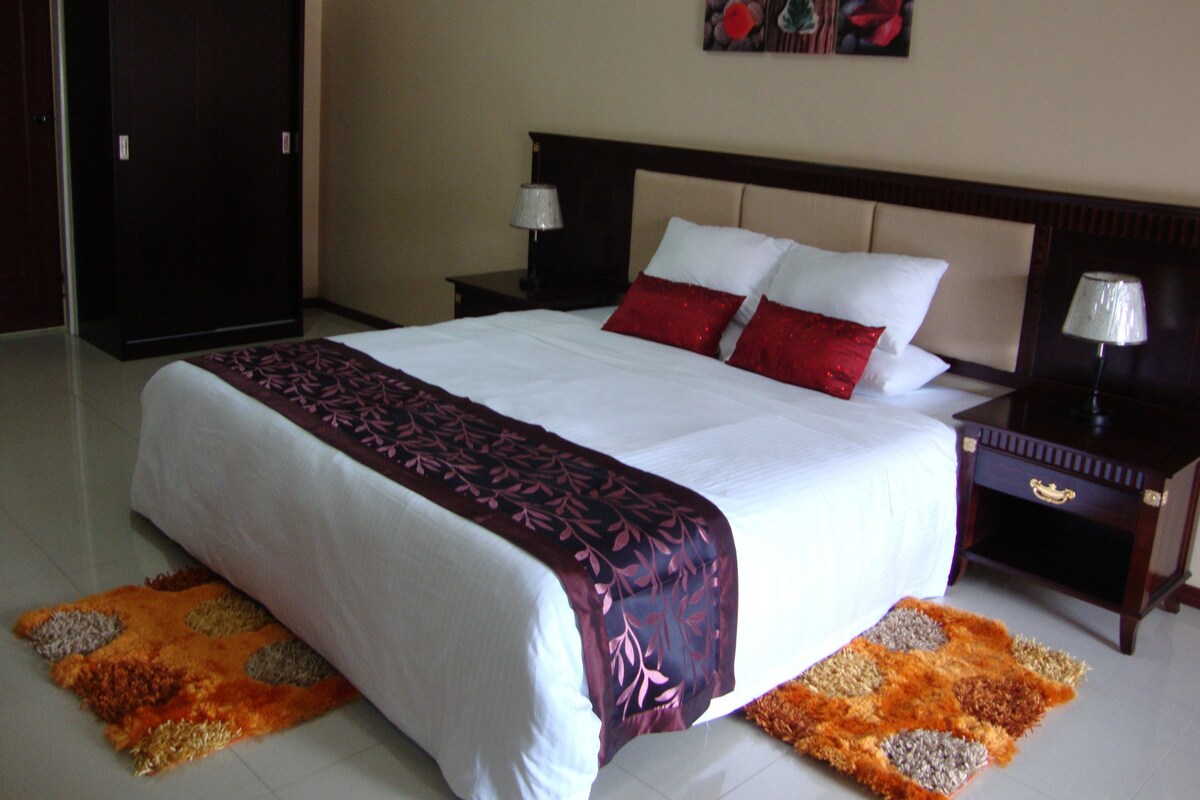 A hotel room with a large bed, two bedside tables, a nightstand, a wardrobe, and a wall-mounted artwork.