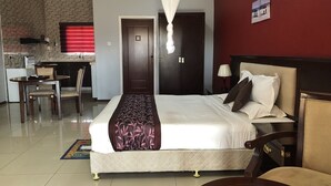 A hotel room with a bed, bedside table, lamp, and a view of the kitchen area. - Riverside Hotel and Conference Centre (Lilongwe)