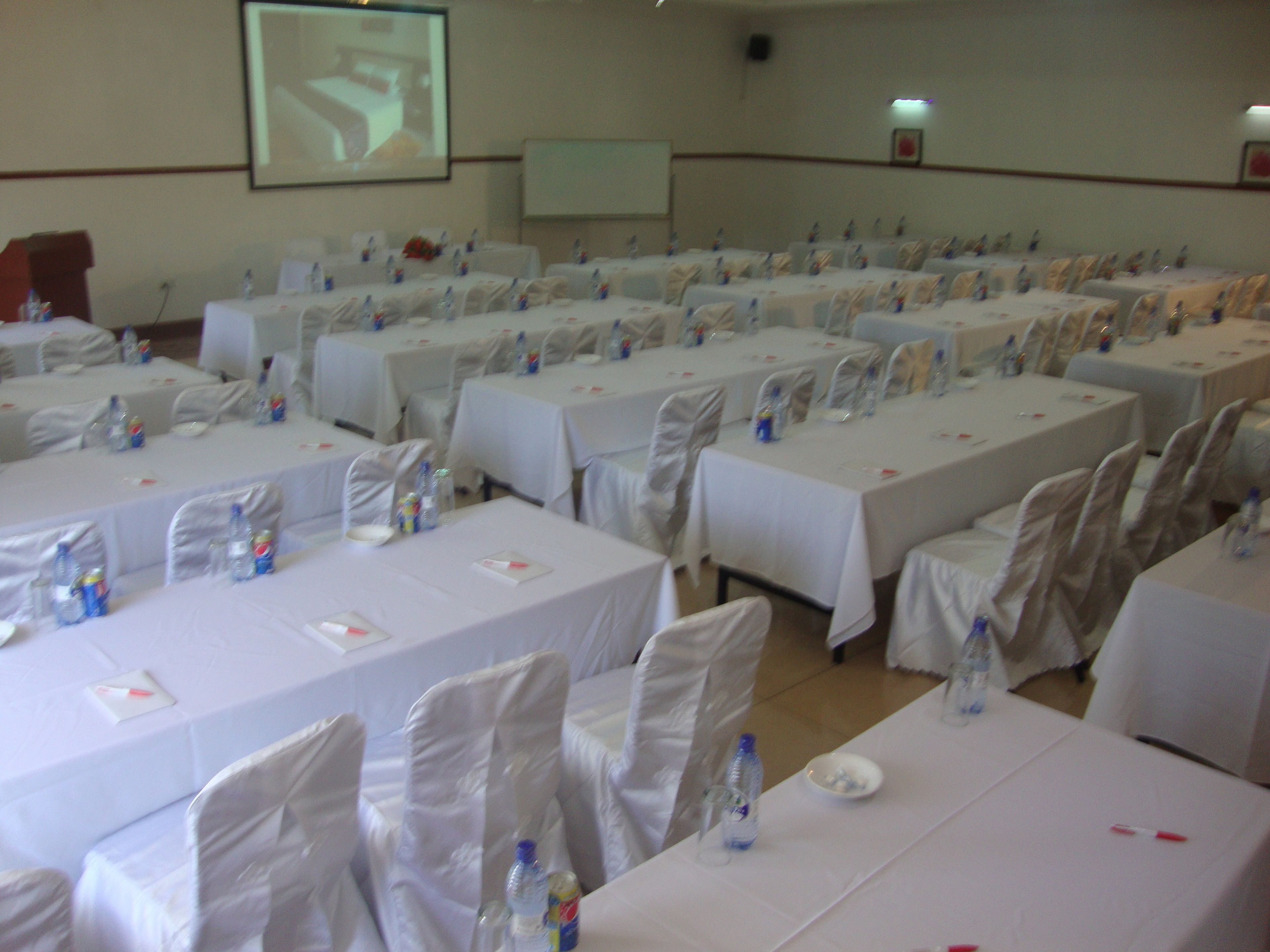 Un salón de conferencias con filas de mesas, cada una con sillas y preparación para banquetear, incluyendo botellas de agua y tarjetas de identificación.