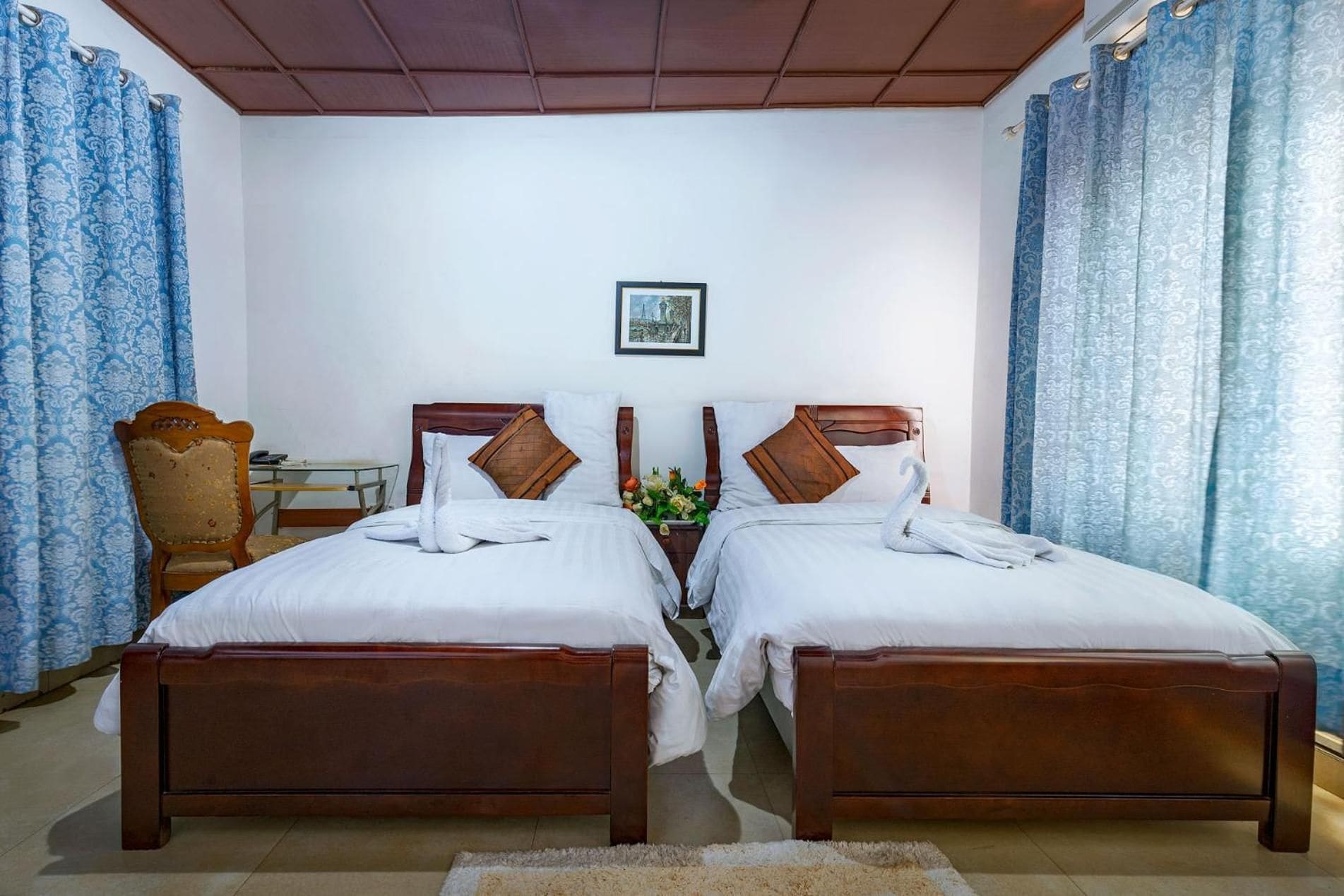 A hotel room with two wooden beds, a wooden chair, a small table, and a framed picture on the wall.