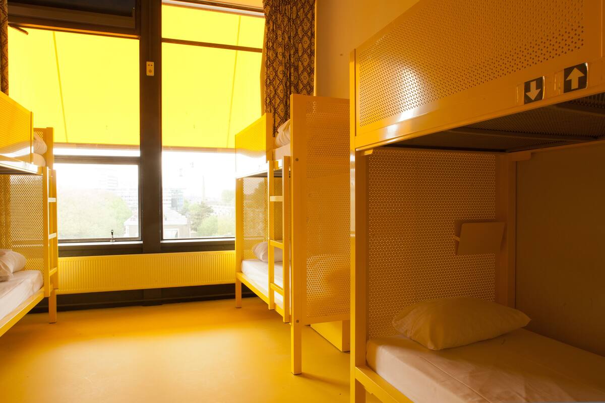 A dormitory room with bunk beds, a window with yellow blinds, and a radiator.