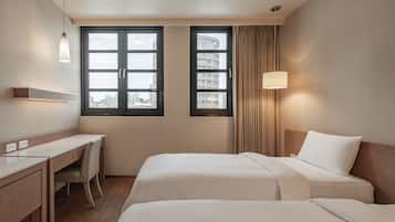 Habitación de hotel con dos camas, un escritorio y una silla. Dos ventanas con cortinas, y una lámpara en la pared.
